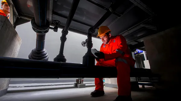 Técnico con casco revisando tuberías mediante inspección de soldaduras y registro digital en espacio reducido.